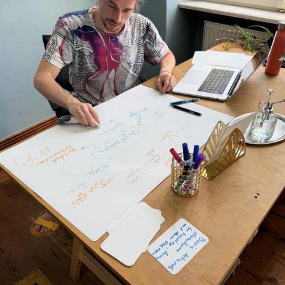 Man at a table writing on a large sheet, with a laptop and a pen holder beside him.