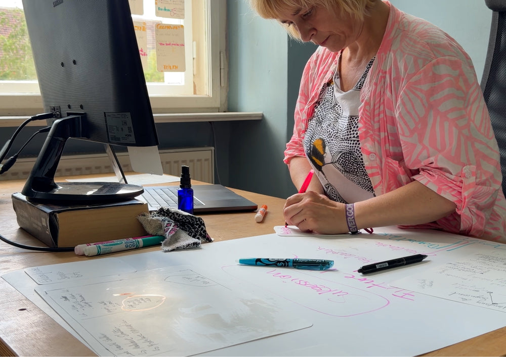 Woman at a desk by the window writing on a large sheet, with a screen and pens beside her.