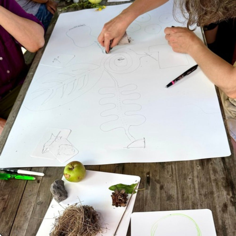 Several hands drawing at the same time on a large sheet on a wooden table outdoors.